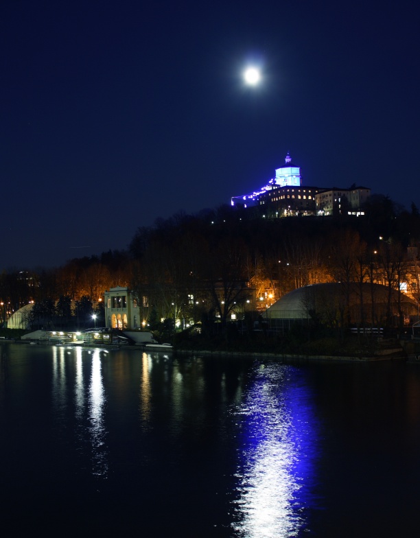 Hinter dem italienischen Fluss Po ragt ein Berg auf, oben steht die Kirche von Turin, darüber leuchtet der Mond, der sich weiß und blau im Wasser spiegelt. 