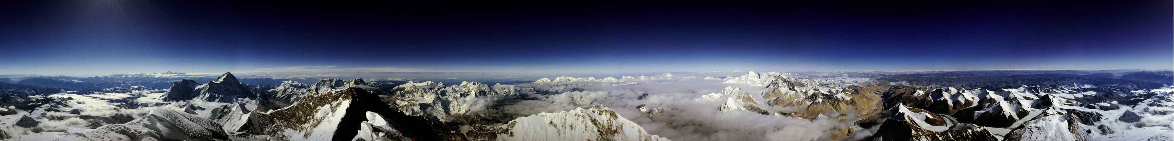 Das sehr breite Pnorama blickt über ein Gebirge, darüber breitet sich ein sehr dunkler Himmel aus.