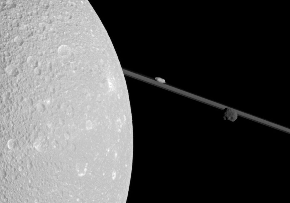 Links füllt der Saturnmond Dione ein Drittel vom Bild. Dione ist von vielen Kratern überzogen, wir sehen nur einen Teil des Mondes. Im Hintergrund verlaufen diagonal die Saturnringe, gespickt von den kleinen Monden Epimetheus und Prometheus.