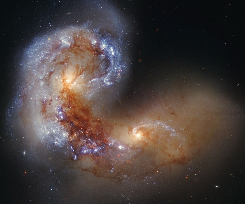 Eine stark verworrene Galaxie wechselwirkt auf diesem Bild mit einer zweiten Galaxie. Die beiden haben hellgelbe Kerne, die linke ist von hellblau leuchtenden jungen Sternhaufen umgeben, vor beiden laufen bräunliche Staubbahnen.