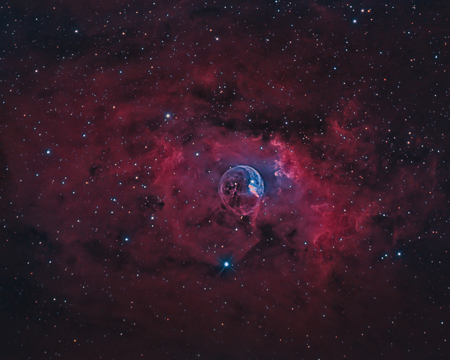Vor einem matten roten Nebelhintergrund leuchtet eine bläuliche Blase, umgeben von roten Ranken. Sie ist als NGC 7635 katalogisiert und auch als Blasennebel bekannt.