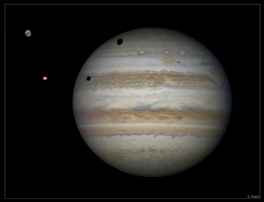 Der Planet Jupiter mit seinen braunen und beigefarbenen Wolkenbändern hat zwei dunkle Flecken. Es sind die Schatten der Monde Ganymed und Io, die links daneben stehe. Am rechten Rand ist der Große Rote Fleck.