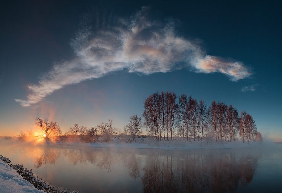 Über dem spiegelglatten Fluss Miass breitet sich am blauen Himmel über einigen Bäumen am Ufer eine Wolke aus. Sie blieb von einem Meteoroid übrig. Links geht die Sonne auf.