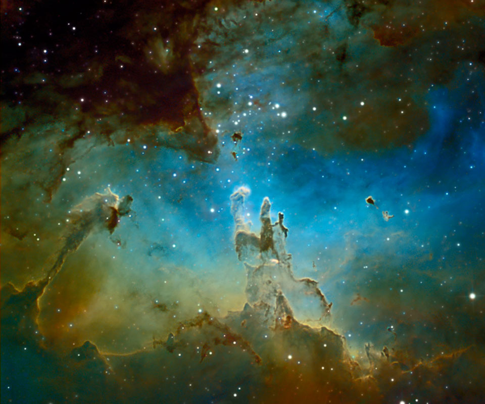 Vor einem leuchtenden Nebelhintergrund, der oben blau und unten gelblich-braun leuchtet, ragt eine dunkle, zerfranste Staubwolke hoch. Es sind die berühmten Säulen der Sternbildung, die auf dem kultigen Hubble-Bild aus dem Jahr 1995 abgelichtet wurden.