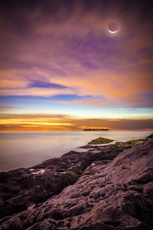 Das Bild zeigt den Strand bei Colonia del Sacramento in Uruguay, dahinter ist die Insel San Gabriel. Am Horizont leuchten die Lichter von Buenos Aires über der Mündung des Rio de la Plata. Hinter zarten Wolken leuchtet der Mond, hinter dem gerade die Venus nach einer Bedeckung wieder zum Vorschein kommt.