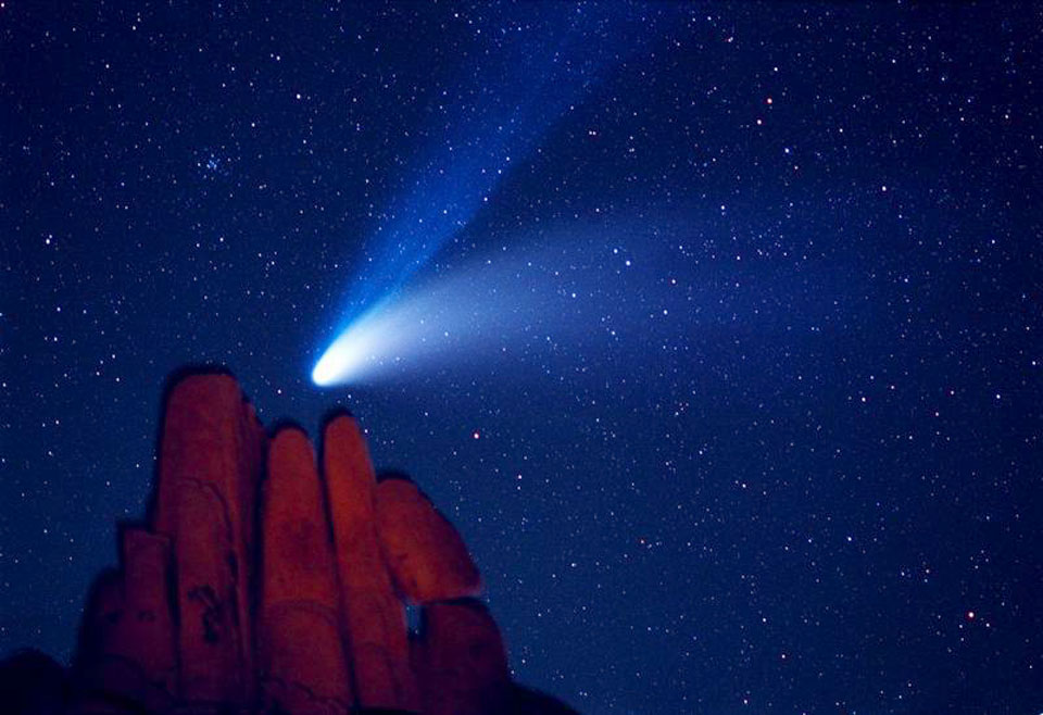 Über roten Felsen, die an Finger erinnern, breitet der Komet Hale-Bopp seine prachtvollen Schweife aus, oben ist der blaue Ionenschweif, darunter der breite weiße Staubschweif. Am Himmel sind leuchten viele Sterne.