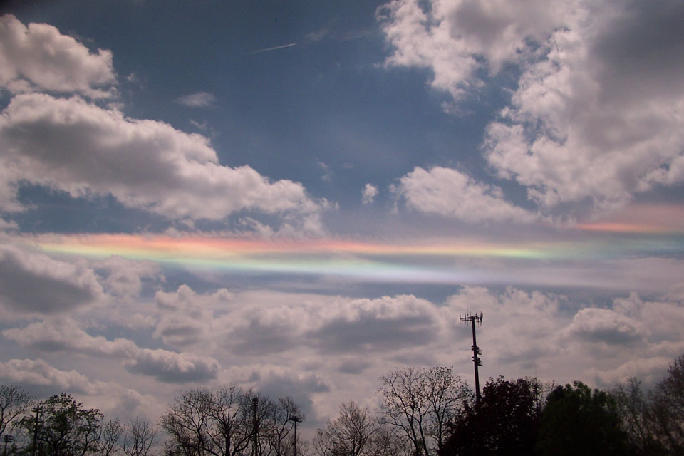 Über den Silhouetten von Bäumen verläuft am wolkigen Himmel ein waagrechter Streifen, der wie ein Regenbogen schillert. Der Himmel wirkt sehr dunkel, weil für das Foto eine Polarisationslinse eingesetzt wurde.