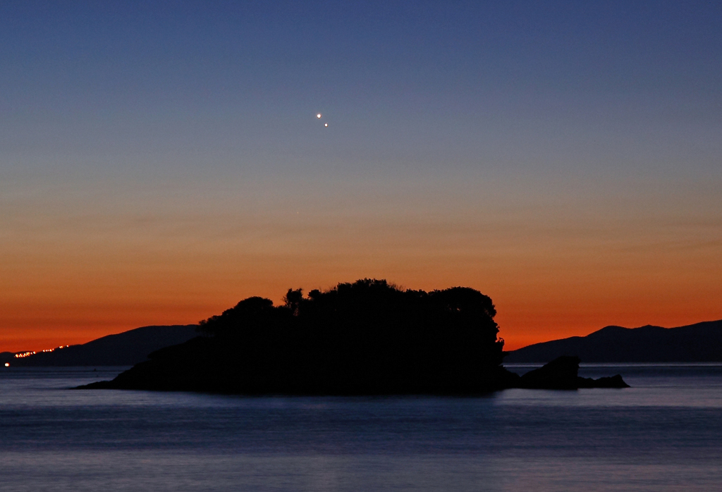 Eine dunkle Silhouette am Horizont ist vorne vom Meer umgeben. Der Himmel verläuft von Dunkelrot nach Tiefblau. Oben glänzen die Planeten Venus und Jupiter eng beisammen.