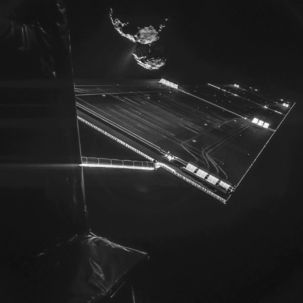 Über einem Solarpaneel der Raumsonde Rosetta schwebt der zweilappige Kern des Kometen 67P/Tschurjumow-Gerassimenko. Von seiner Oberfläche strömt ein weißer Strahl aus.