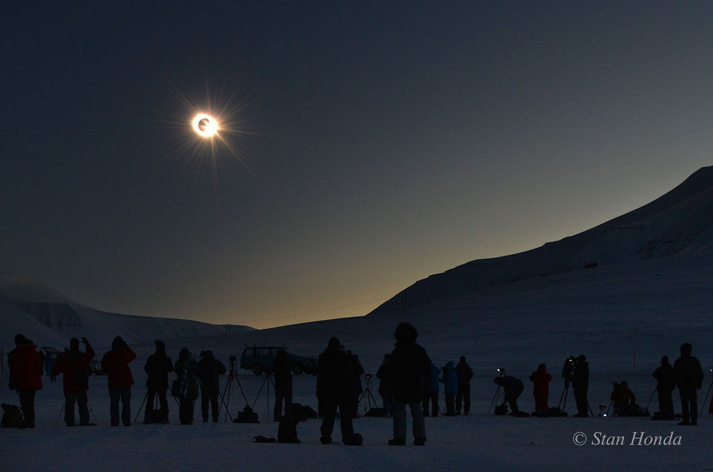 Am dunklen, kristallklaren Himmel einer verschneiten Landschaft gibt der Mond gerade wieder die Sonne frei. Am Mondrand blitzt ein gleißender Diamantring. Vorne stehen viele dunkle Silhouetten von Finsternisjägerinnen*.