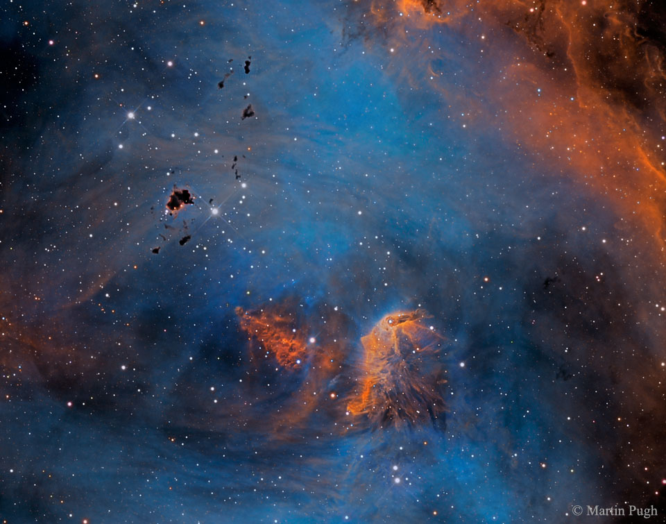 Vor einem blauen Hintergrund schweben einige orange Nebel und dunkle Wolkenfetzen aus dichtem Staub, links unten sind mehr Sterne als rechts oben, insgesamt sind relativ wenige Sterne im Bild. Die Region heißt auch Collinder 249.