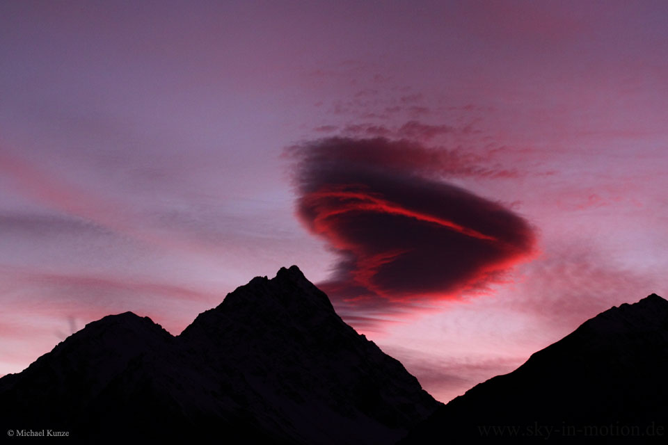 Über den dunklen Silhouetten von Bergen schwebt eine herzförmige rote Wolke. Es ist eine seltsam geformte Lenticularis.