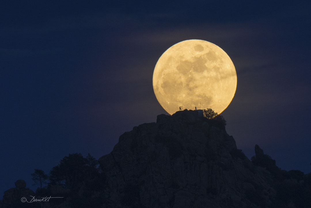 Über einem Berggipfel schwebt der Mond, er hat ein Gesicht. Auf dem Berg steht eine Frau mit Teleskop. Sie zeichnet sich als Silhouette vor dem Mond ab.