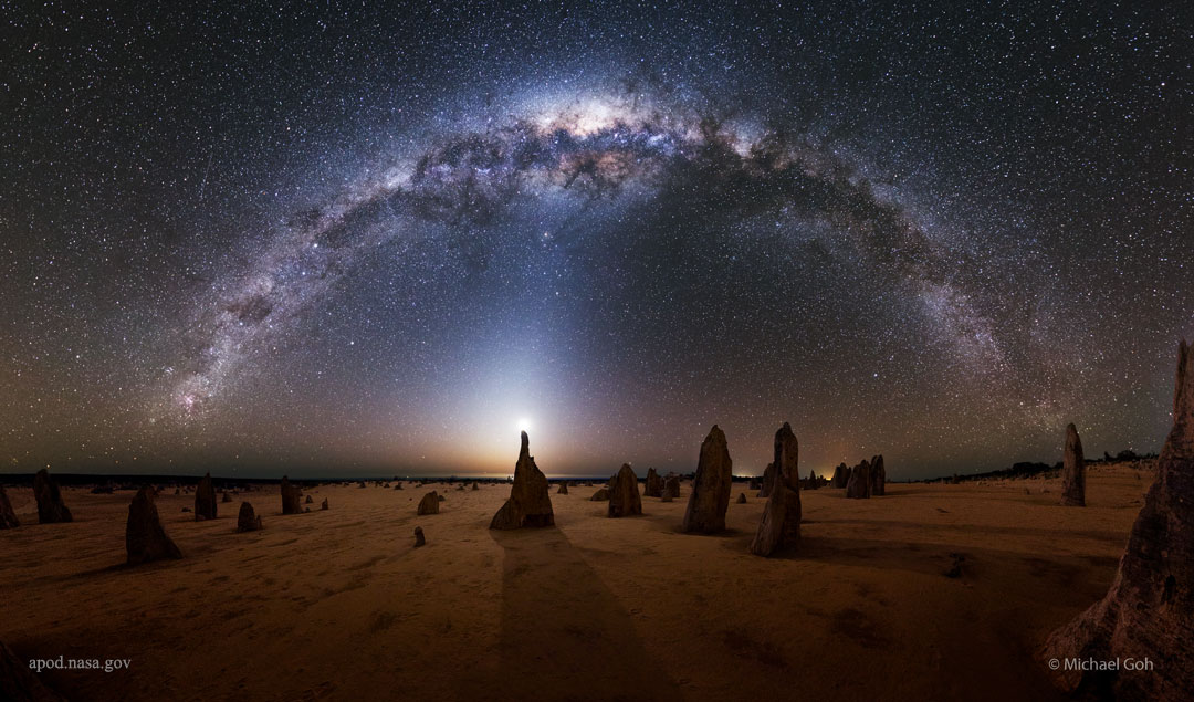 Aus einer mondbeleuchteten flachen Landschaft im Nambung-Nationalpark ragen spitze Felsen aus Kalkstein auf, es sind sogenannte Pinnacles. Hinter einer Spitze strahlt der Mond. Die Milchstraße wölbt sich in einem Bogen im Bild. Vom Mond steigt zartes Zodiakallicht auf.