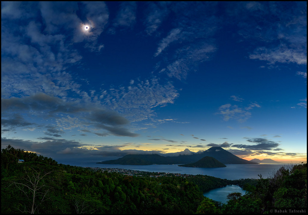 Der Himmel über Vulkankegeln, die aus dem Meer ragen, ist dunkelblau und von Wolken überzogen. Am Horizont ist ein heller Rand. Links oben ist die dunkle Silhouette des Mondes von der hellen Sonnenkorona umgeben.