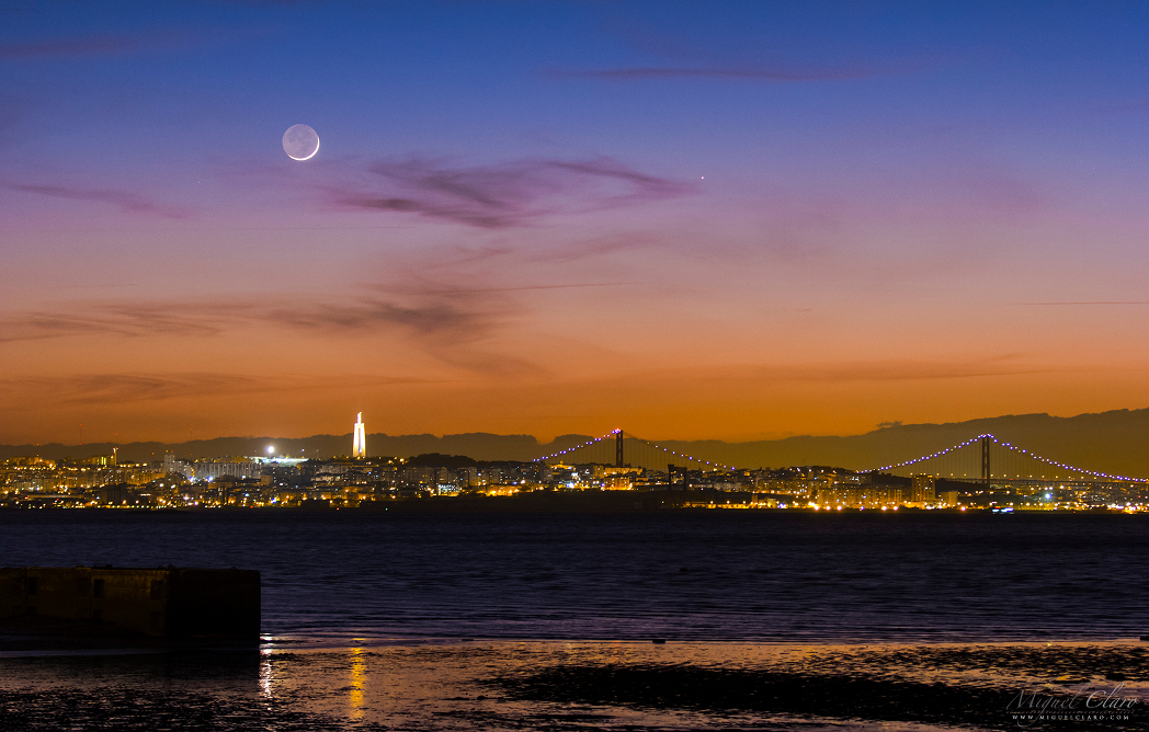 Über der Hafenstadt Lissabon und dem Tejo leuchten der Sichelmond und der Planet Merkur nach Sonnenuntergang im Abendrot.