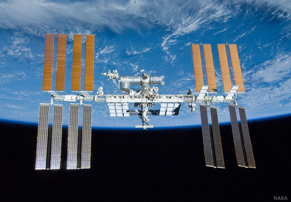 Von oben ragt die Erde ins Bild, darunter ist der schwarze Weltraum. Vor dem Horizont schwebt die Internationale Raumstation mit ihren ausladenden Solarpaneelen und den Modulen, in denen Menschen forschen.