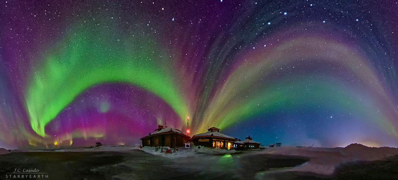 Die Polarlichter wirken wie Bögen, weil das Panorama 360 Grad breit ist. Die Bögen wölben sich von einem Haus ausgehend nach links und rechts.
