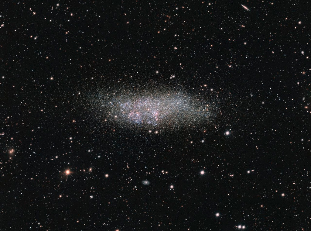 Mitten im Bild schwebt die kleine Galaxie WLM im Sternbild Walfisch. Sie wirkt unregelmäßig und ist von rosaroten Sternbildungsregionen und blauen Sternen gesprenkelt.