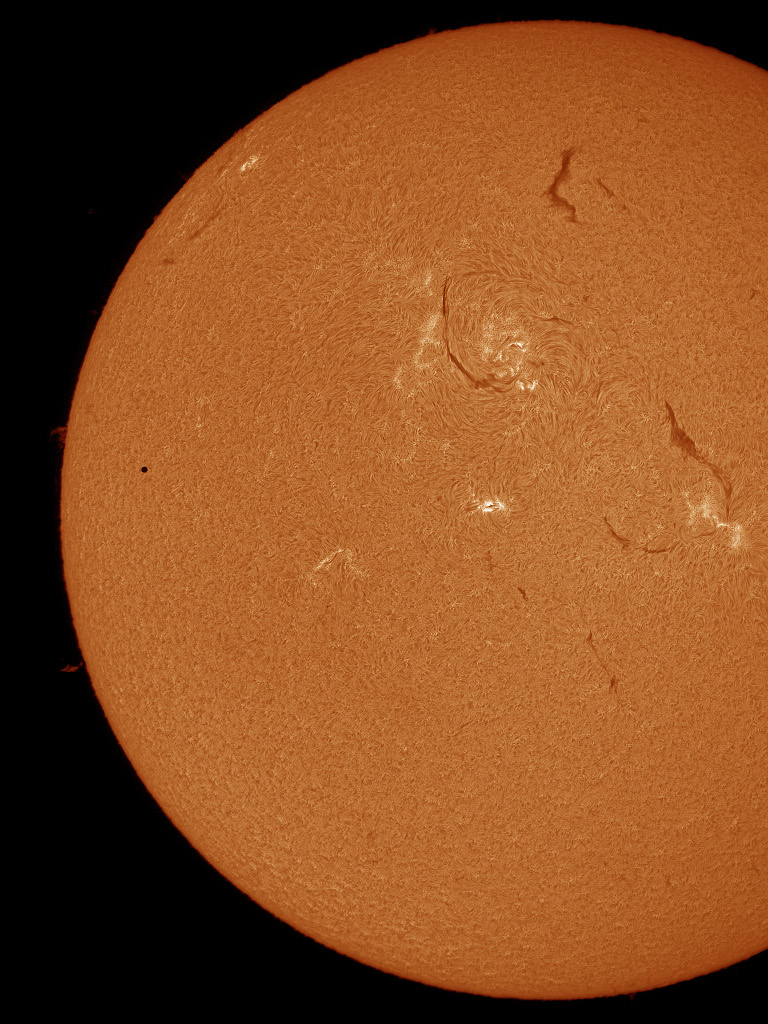 Die orangefarbene Sonne ist teilweise abgebildet, links und unten sieht man ihren Rand. Beim rechten Rand steht der Planet Merkur, er ist ein winziger schwarzer Kreis. Die Sonne ist von dunklen und hellen Fäden überzogen.