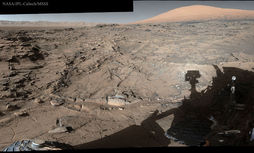 Der Schatten des Marsrovers Curiosity fällt auf zerklüftetes Gelände. Hinten am Horizont ragt rechts der Mount Sharp auf (Aeolis Mons), er ist das Ziel des Rovers.