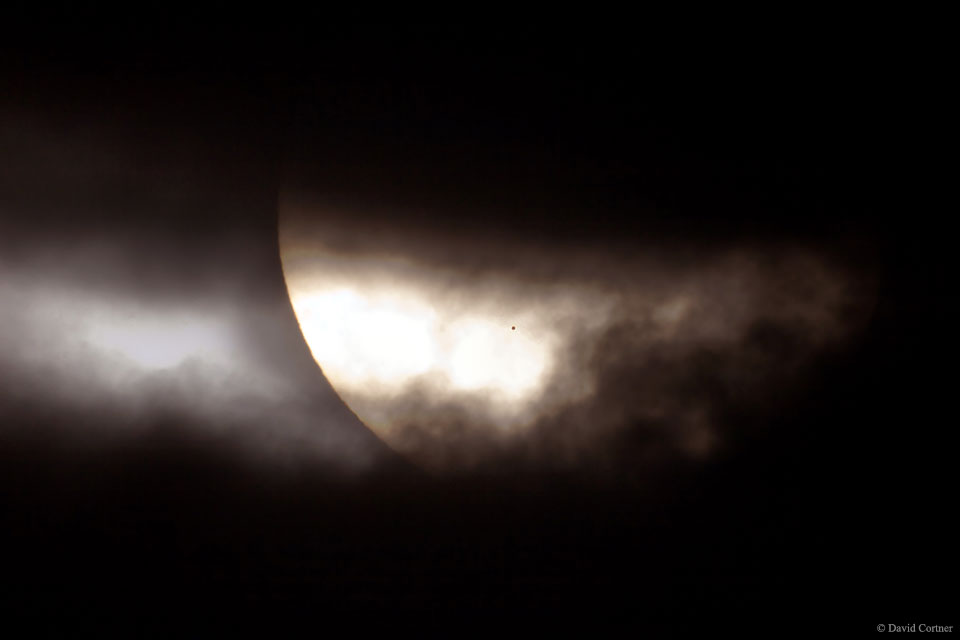 Die Sonne schaut zwischen dunklen Wolken heraus. Man sieht nur einen kleinen Teil davon. Rechts unter einer Wolke ist ein winziger kreisrunder schwarzer Fleck. Es ist der Planet Merkur bei einem Merkurtransit.