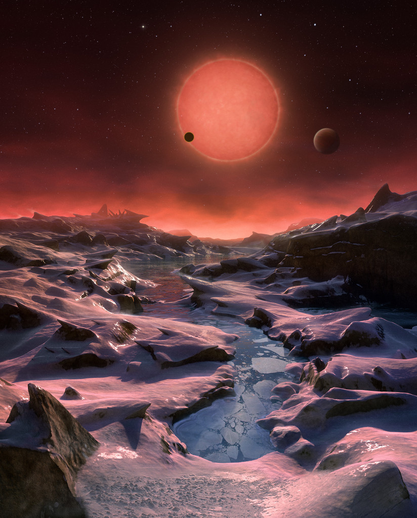 Diese Illustration zeigt den Horizont eines Planeten, der um den Zwergstern TRAPPIST-1 kreist. Ein Planet zieht gerade vor dem roten Stern vorbei, ein anderer steht etwas größer rechts neben dem Stern. Die Landschaft ist schroff, vorne ist ein Gewässer mit Eisschollen, der Himmel wirkt düster und dämmrig, oben sind aber Sterne erkennbar.