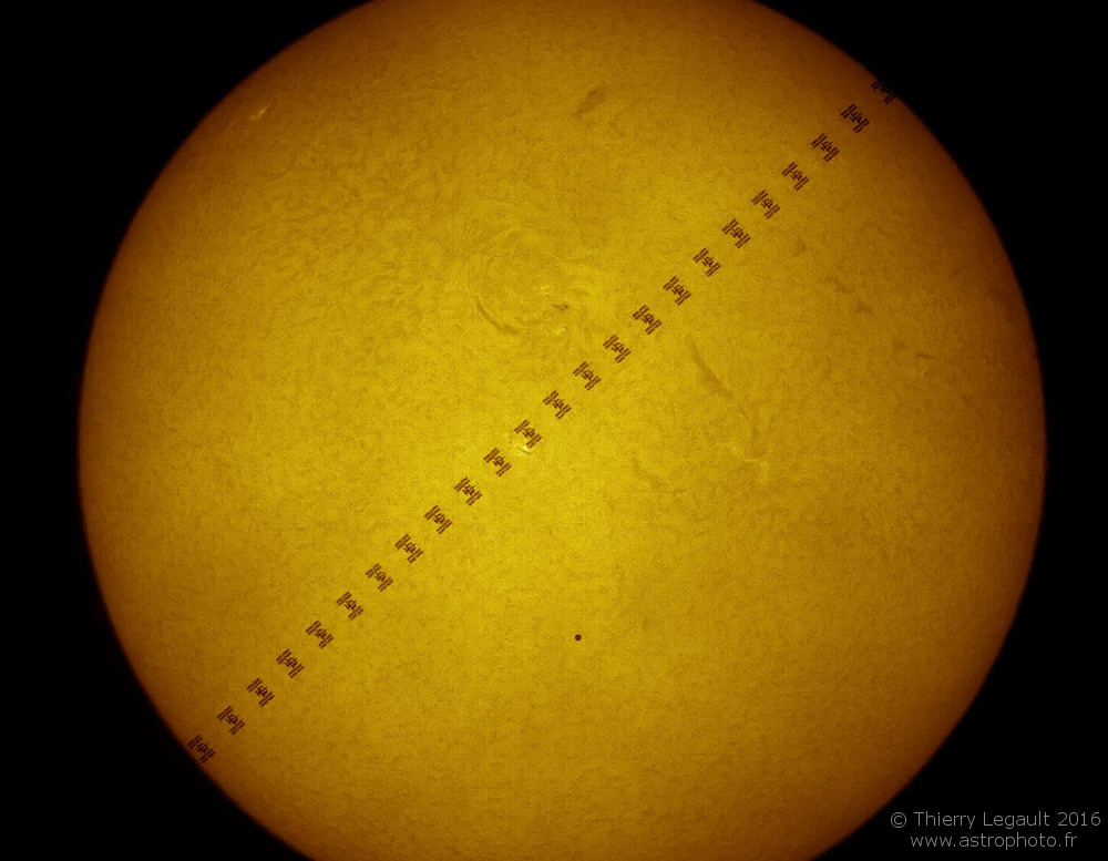 Auf der orange-gelben Sonne zeichnen sich Silhouetten ab. Diagonal zieht die Internationale Raumstation mit ihren Paneelen über die Sonne, unter der Mitte ist ein unscheinbarer, kleiner, dunkler Punkt, es ist der Planet Merkur bei einem Merkurtransit.