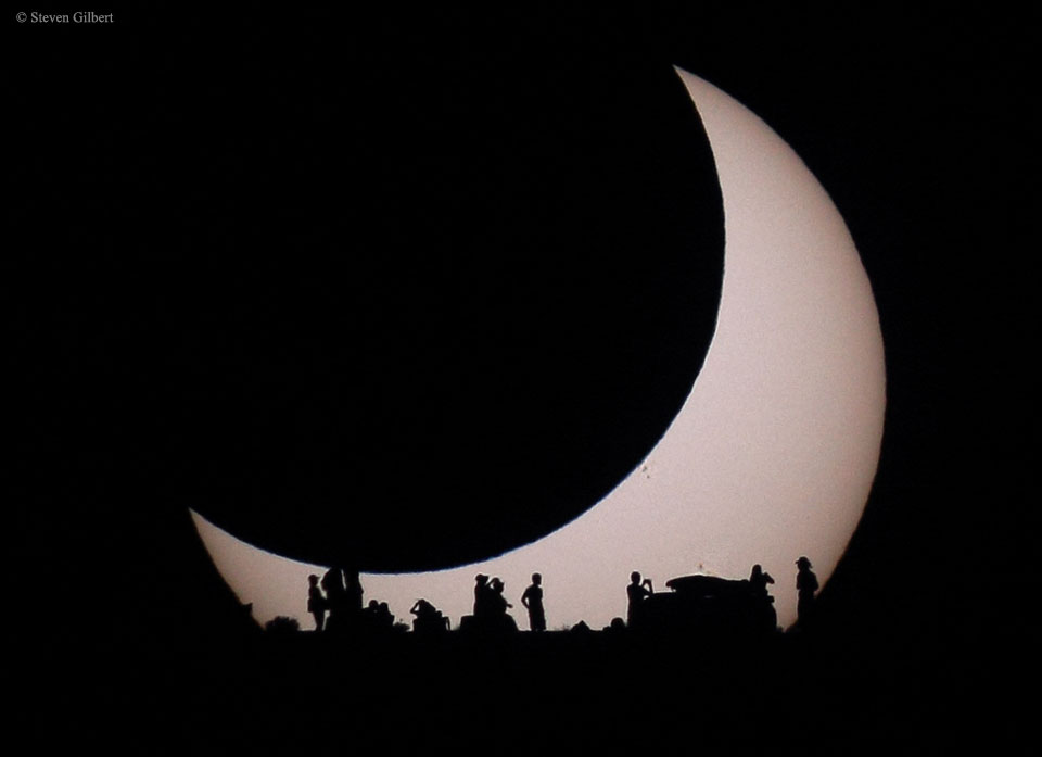 Der Mond bedeckt die Sonne, während sie aufgeht. Am Horizont sind die Silhouetten von Beobachtenden vor der Sonne zu sehen. Auch Sonnenflecken verdunkeln den Zentralstern im Sonnensystem.