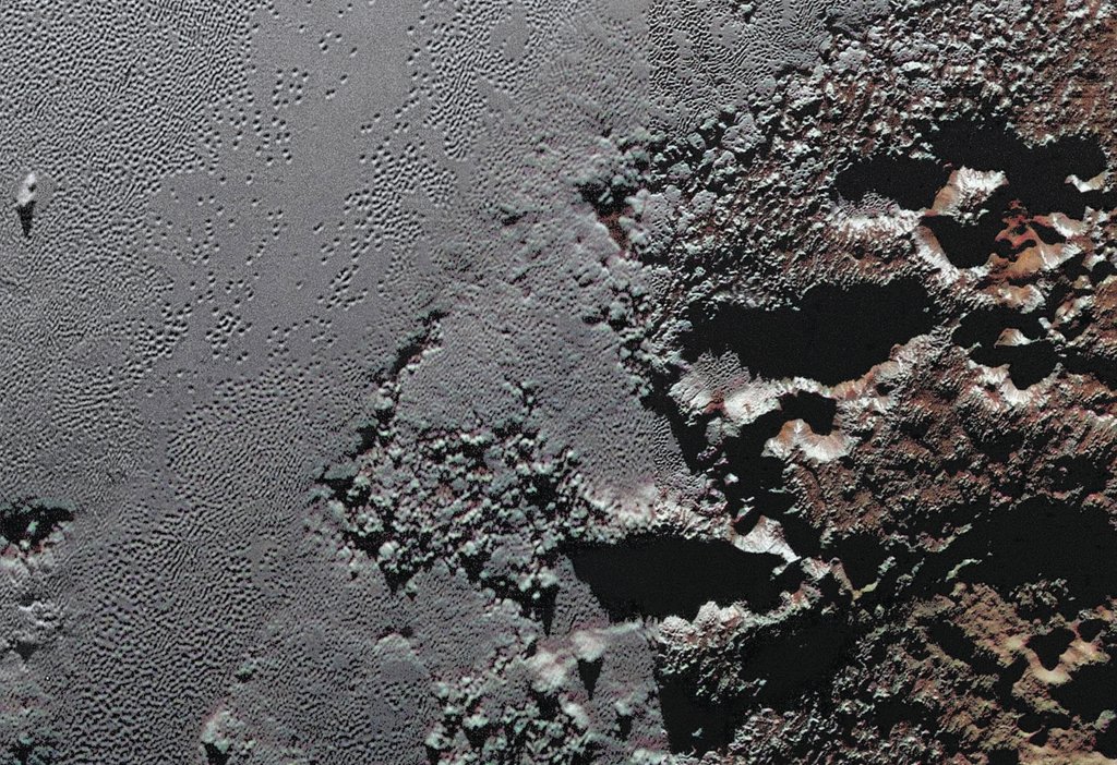Links ist ebenes, helles Gelände, das von Gruben bedeckt ist. Rechts ragen hohe Berge auf, die rötlich gefärbt sind. Das exotische Gelände befindet sich auf dem Zwergplaneten Pluto, es wurde von der Raumsonde New Horizons abgebildet.