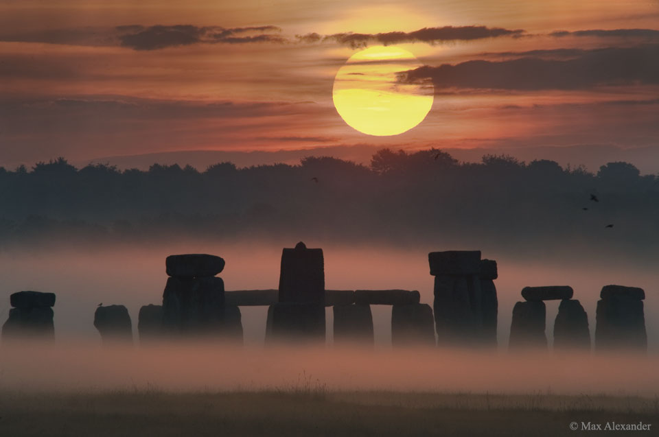 Aus dem Bodennebel ragen die Monolithe von Stonehenge auf. Darüber geht die Sonne am wolkigen Himmel auf. Das Bild vom Sonnenaufgang ist orange gefärbt.