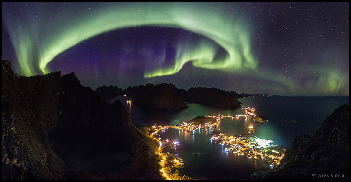 Polarlichter über Reine auf Lofoten in Norwegen. Die Küste ist hell erleuchtet und zeigt, dass sogar entlegene Gebiete mit Lichtverschmutzung kämpfen.