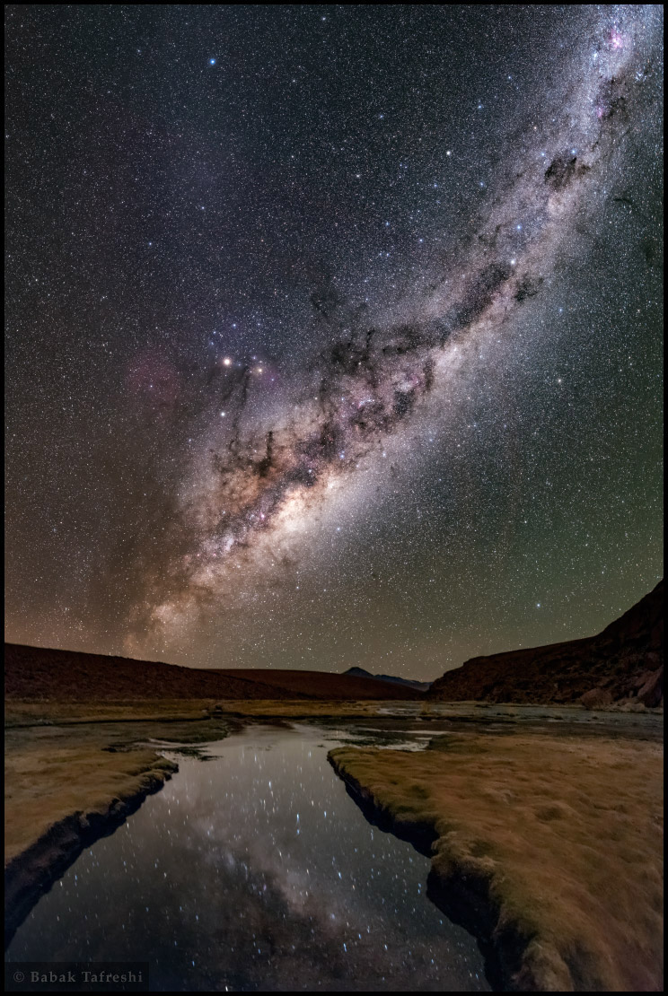 Über dem Altiplano steigt die Milchstraße auf. Sie spiegelt sich vorne in einem ruhigen Fluss. Hinten steht ein Vulkan am Horizont. Über einem Dreieck aus Mars, Saturn und Antares steigt Zodiakallicht auf. Links daneben schimmert der riesige rote Nebel Sh 2-27.
