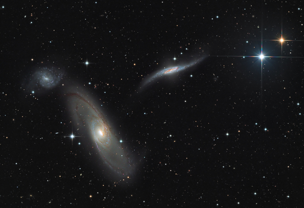 Drei Galaxien links im Bild sind ein Trio, sie wechselwirken miteinander. Rechts oben leuchten ein blauer und ein gelber Stern, beide haben Zacken und liegen in der Milchstraße.