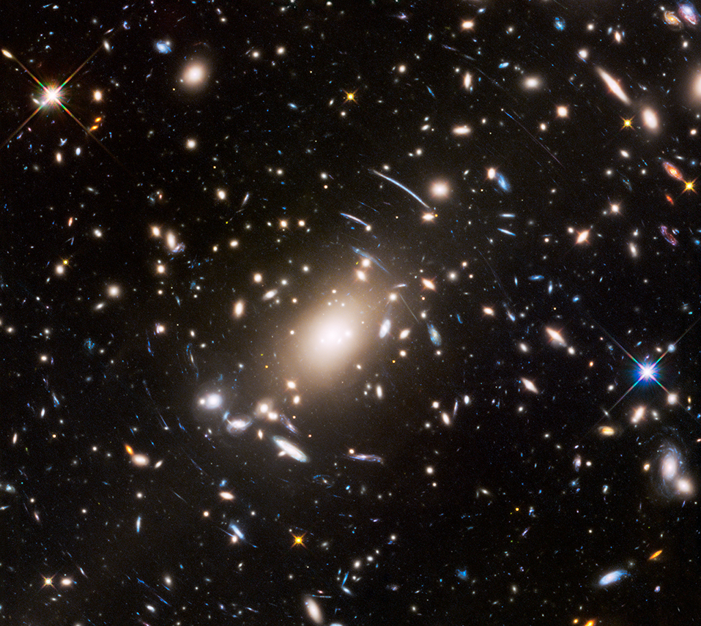 Zwischen den Galaxien des Haufens Abell S1063 im Sternbild Kranich verlaufen gekrümmte blaue Bögen. Es sind Bilder von Galaxien, die viel weiter entfernt sind. Die Bögen entstehen durch den Gravitationslinseneffekt.