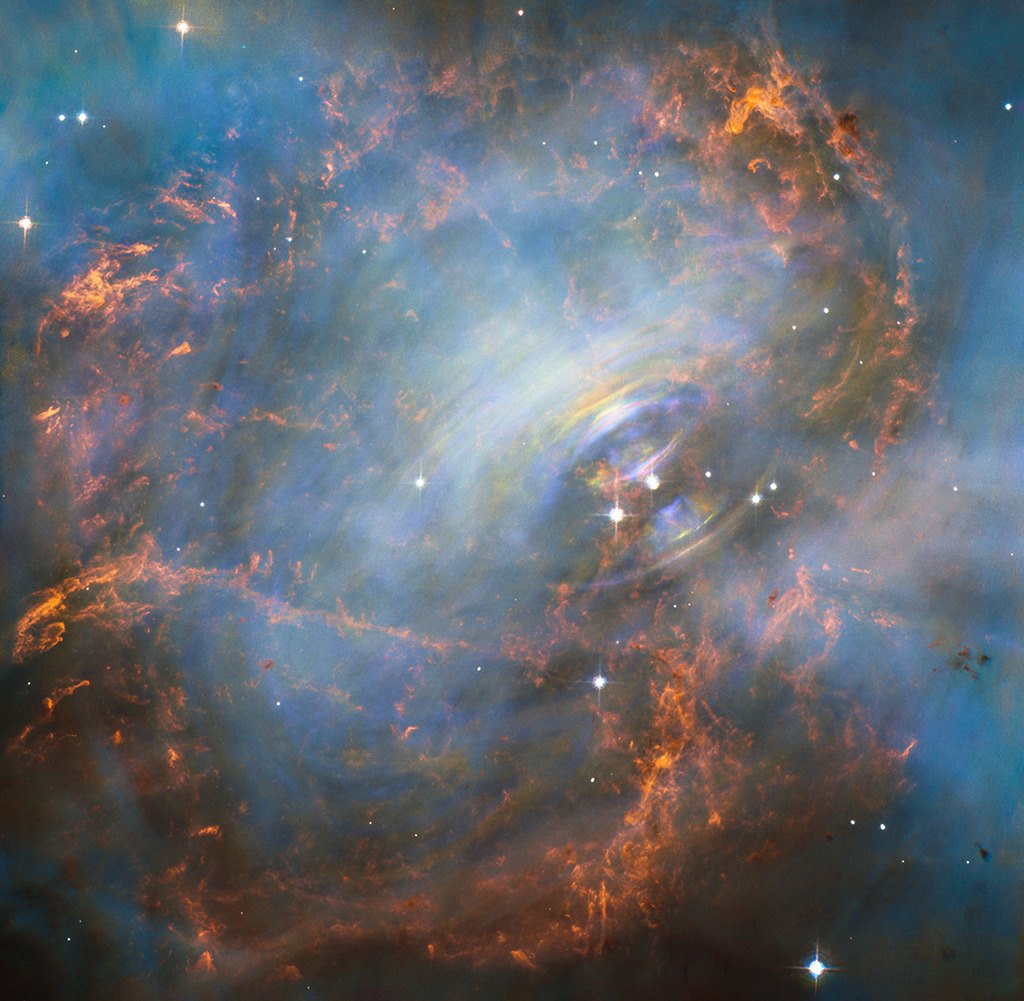 Hinter weißlichen Nebeln leuchten rote Fasern und blaue Nebel. Rechts neben der Mitte sind spiralförmige Wirbel zu sehen. Dort befindet sich auch der Krebspulsar, der nach der Explosion eines massereichen Sterns übrig blieb.