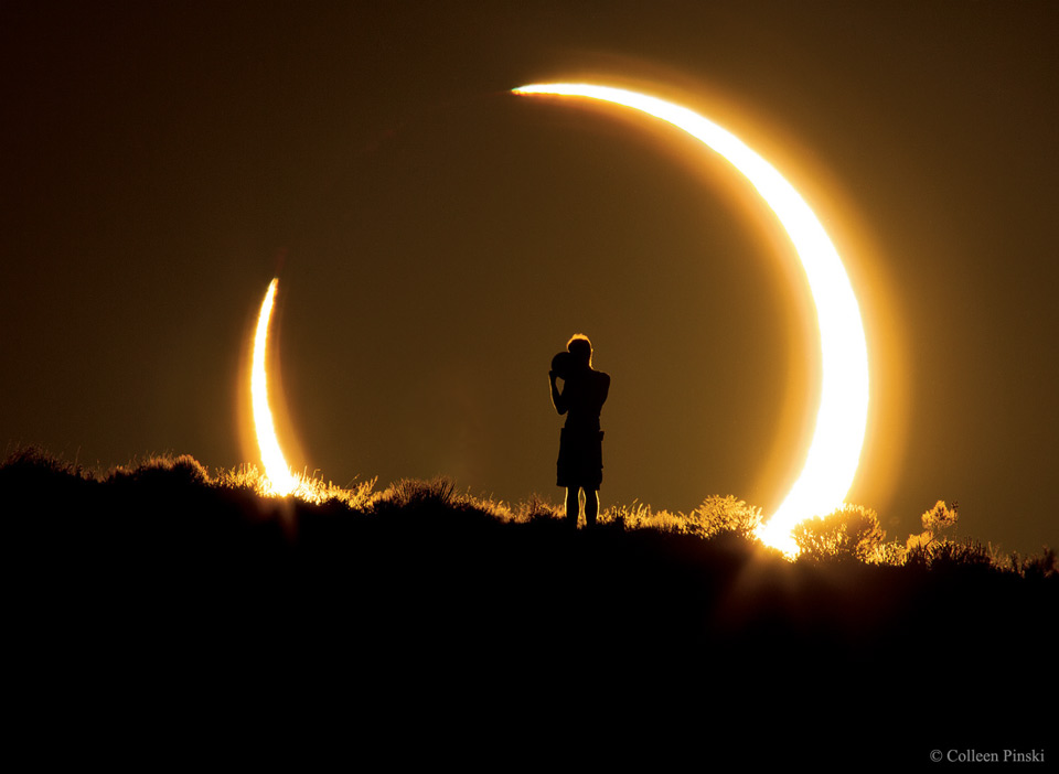 Über einem Hügel geht die Sonne unter. Sie sieht aus wie eine Sichel, weil der Mond sie teilweise bedeckt. Mitten vor der ringförmigen Sonnenfinsternis steht eine Person, die ein Gerät zur Sonne hält.