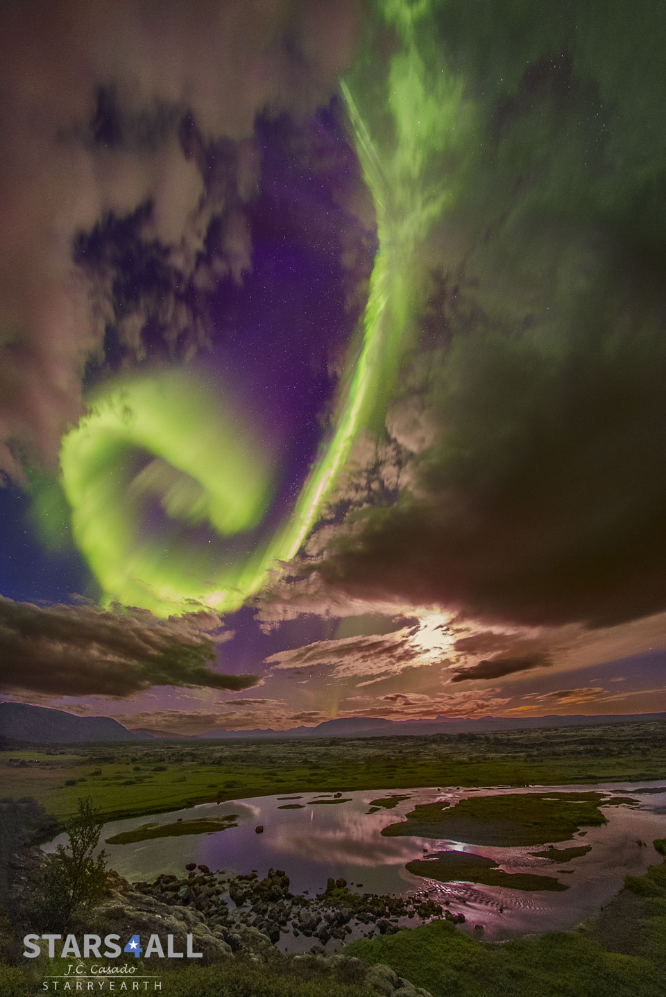Über dem isländischen See Þingvallavatn ringelt sich ein spiralförmiges Polarlicht von oben ein. Rechts daneben schimmert der Mond hinter Wolken.