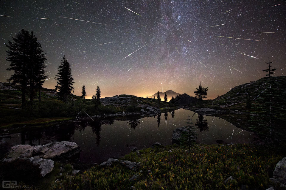 Hinter einem ruhigen Bergsee ragt ein ferner Gipfel am Horizont auf. Darüber zischen Meteore über den Himmel, an dem die Milchstraße senkrecht aufsteigt.