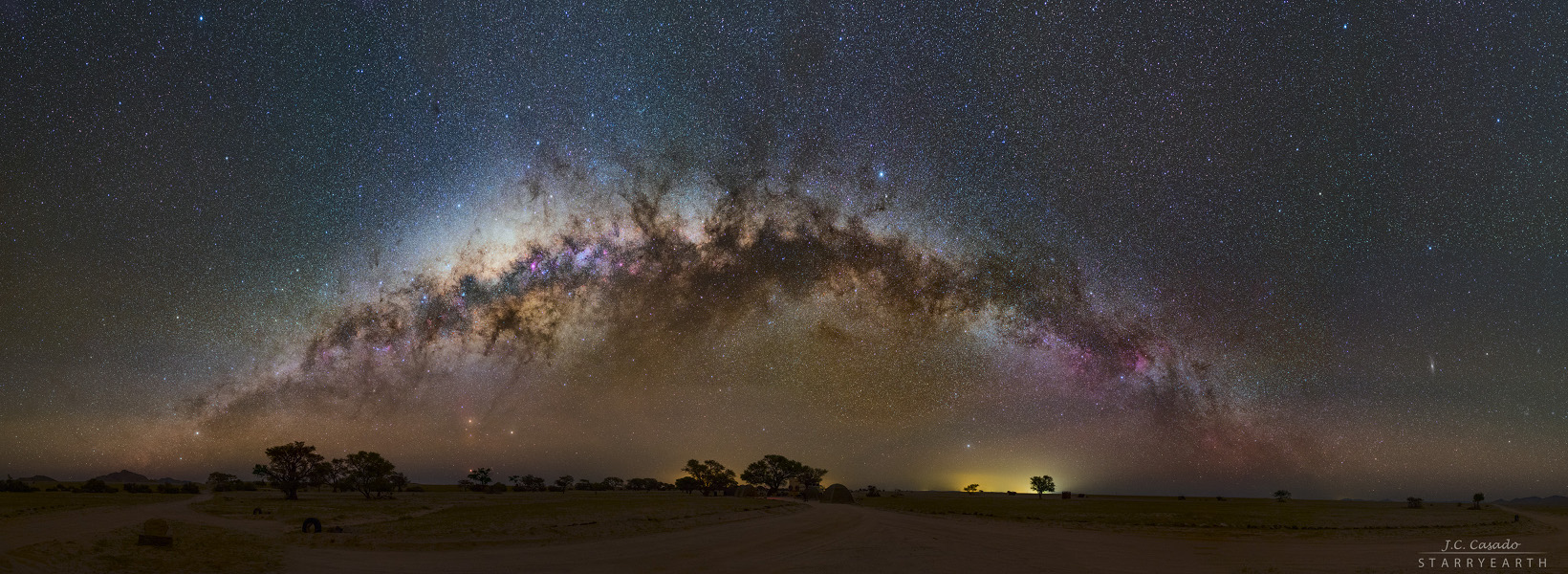 Über dem Horizont wölbt sich die Milchstraße mit diffusen Sternwolken und dunklen Staubwolken. Darunter bilden die Planeten Mars und Saturn mit dem hellen Stern Antares ein Dreieck knapp über den Bäumen.
