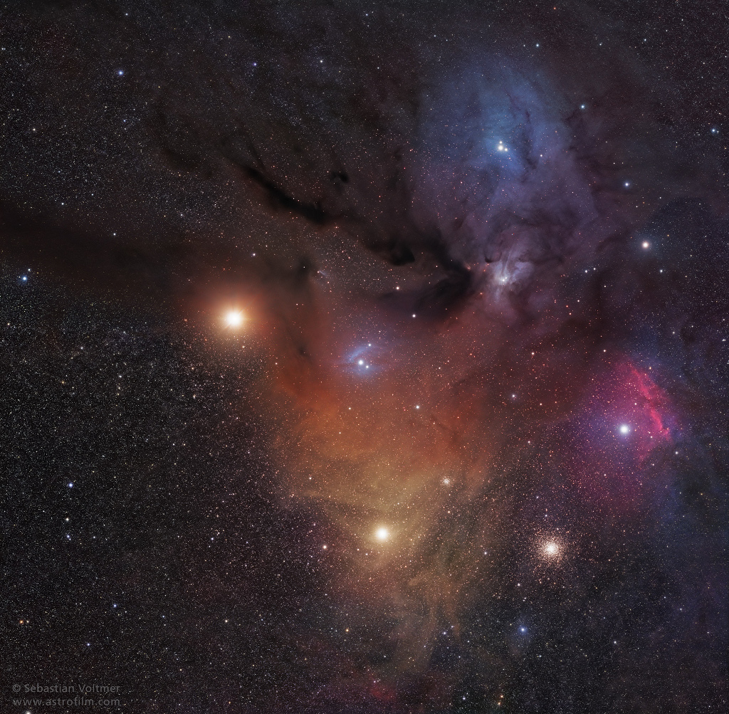 Zwischen bunten Nebeln sind helle Lichter verteilt, im Hintergrund sind Wolken aus zarten Sternen. Eins der hellen Lichter ist ein Kugelsternhaufen. Die Nebel sind Reflexionsnebel und Emissionsnebel. Auch der Planet Mars ist im Bild.