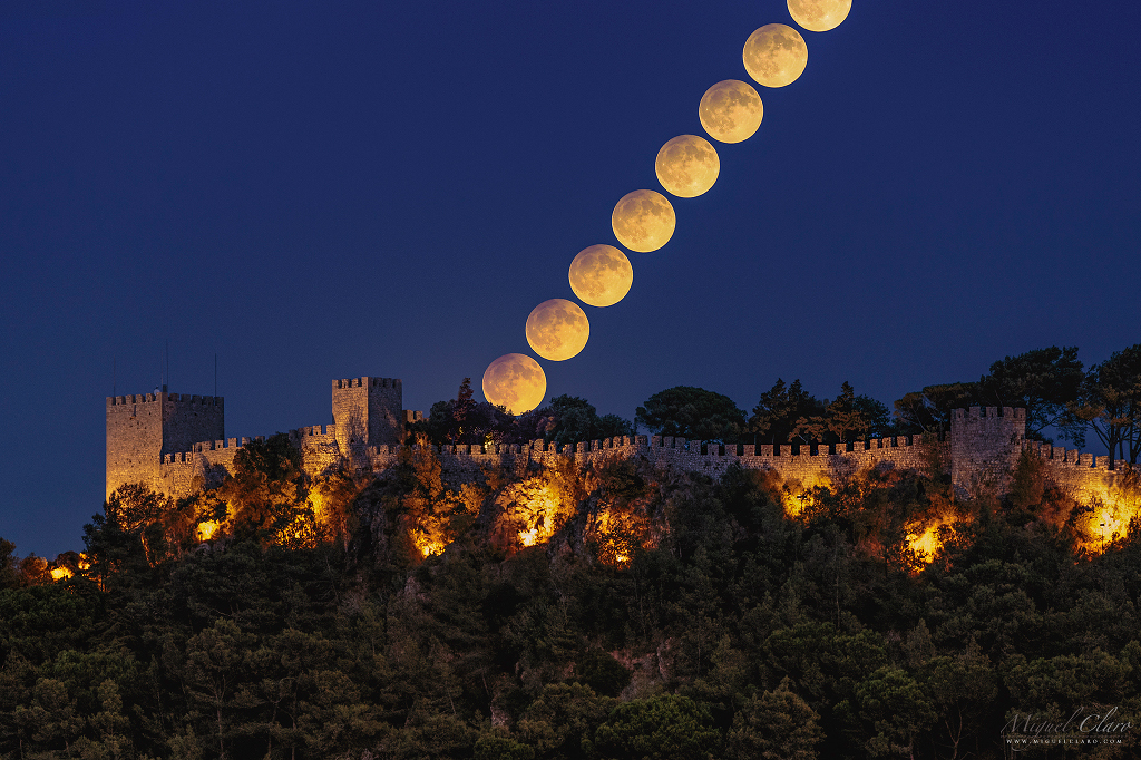 Über den beleuchteten Festungsmauern des Castelo de Sesimbra geht der volle Erntemond am dunkelblauen Himmel auf. Der obere Mondrand wird von einer Halbschattenfinsternis gedimmt.