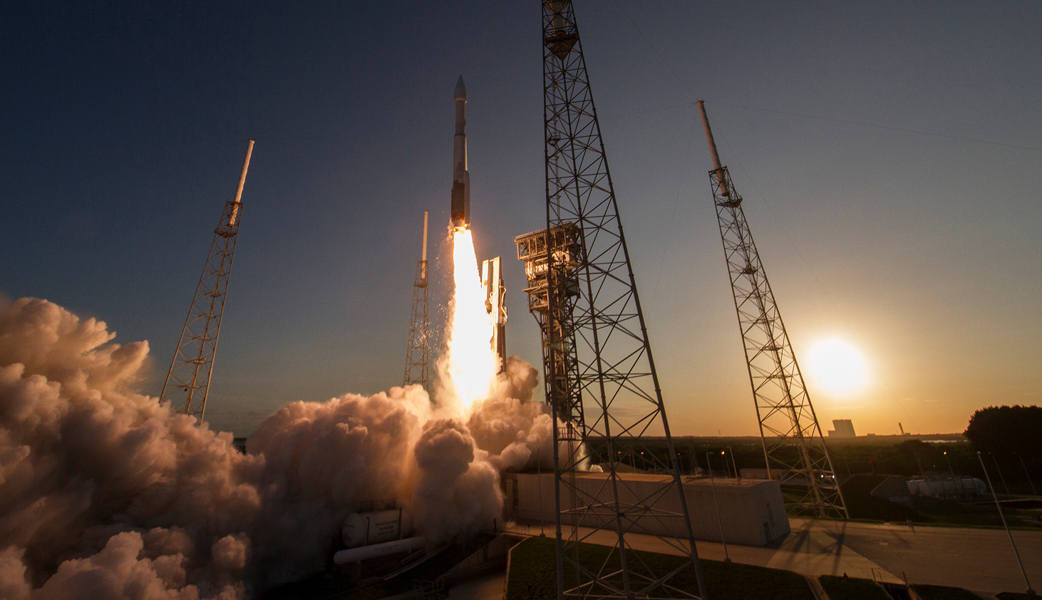 Die Szene ist orangefarben beleuchtet. Rechts ist die Sonne, links der Feuerstrahl einer Rakete, die gerade startet. Sie ist von gerippeförmigen Türmen umgeben.
