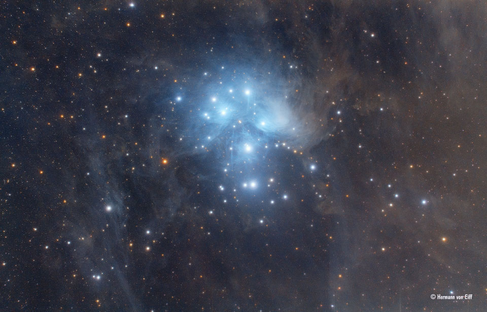 Der kompakte Sternhaufen der Plejaden (M45) ist von dichten Staubwolken umgeben. In der Nähe der Sterne sind sie bläulich beleuchtet. Außen herum sind weitere dunkle Staubwolken.