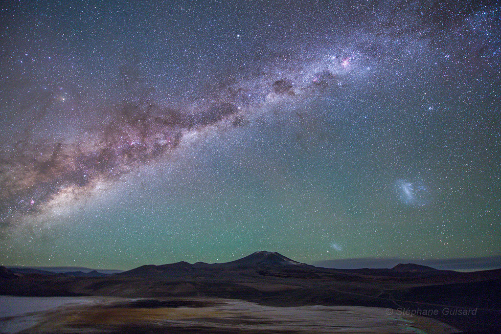 Über der Altiplano in der Atacama im Norden von Chile geht die Milchstraße auf. Darunter schimmern die Magellanschen Wolken. Am Horizont ragt der Schichtvulkan Lastarria auf. Die ruhige Szene wirkt fast außerirdisch.