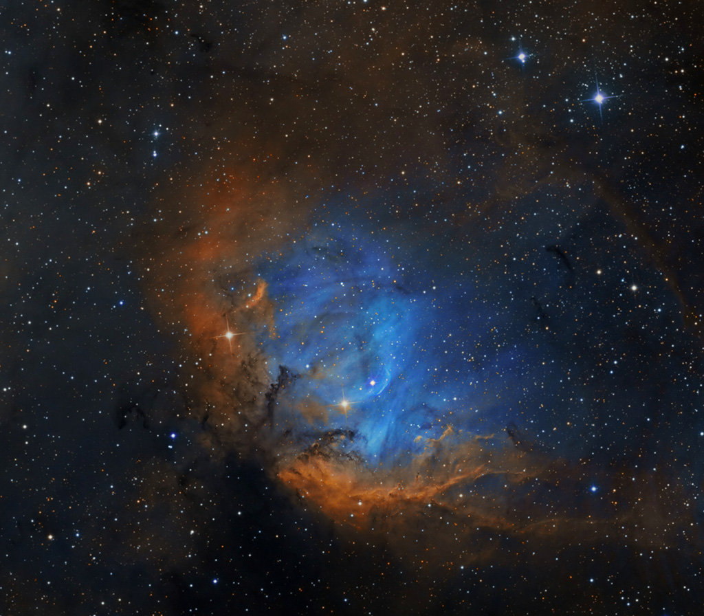 Ein orangefarbener Nebel ist schalenförmig nach rechts geöffnet, darin schimmert ein blauer, gefaserter Nebel, links unten sind zwei kleine dunkle Staubranken. Außen herum sind viele kleine Sterne gleichmäßig verteilt.