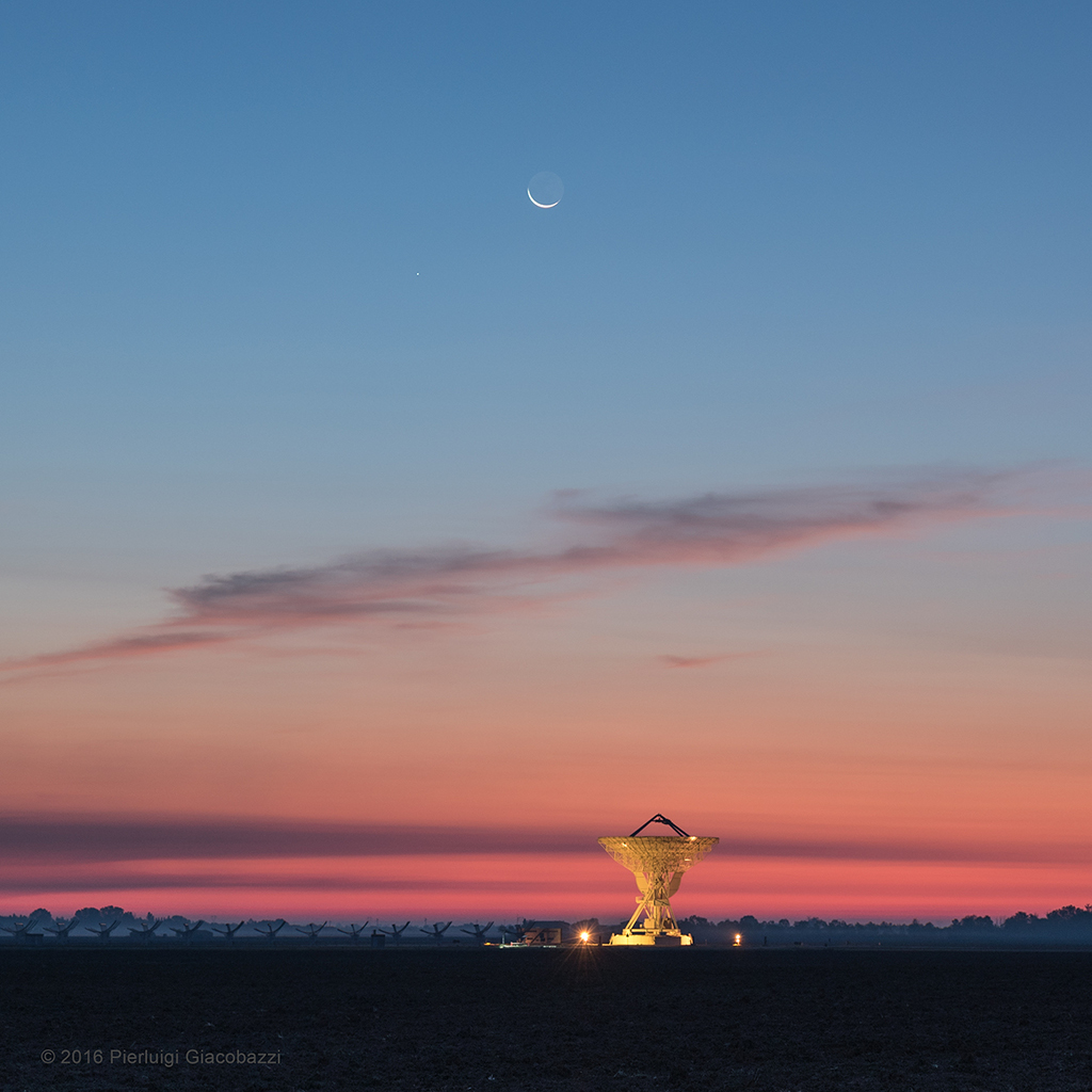 In der Abenddämmerung steht eine gelb beleuchtete Radio-Parabolantenne, die Schüssel ist nach oben gerichtet. Darüber verfärbt sich der Himmel von leuchtend rot aufwärts nach mittelblau. Oben steht der Mond, die schmale Sichel zeigt nach unten, die Nachtseite nach oben. Links unter dem Mond leuchtet Merkur.