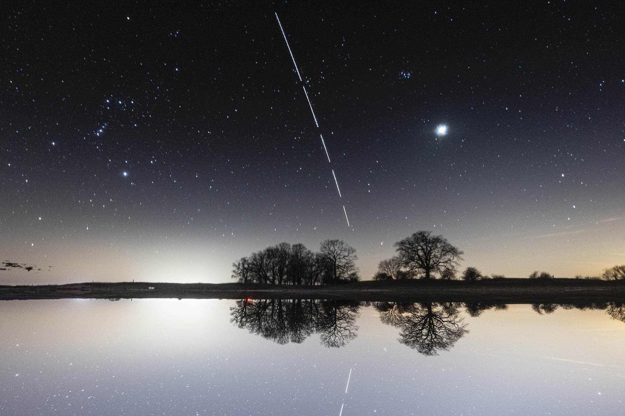 Die Internationale Raumstation ISS zieht über den klaren Abendhimmel mit Orion, Venus und den Plejaden über einer Wasserstelle in Wulfsahl bei Gusborn in Norddeutschland.
