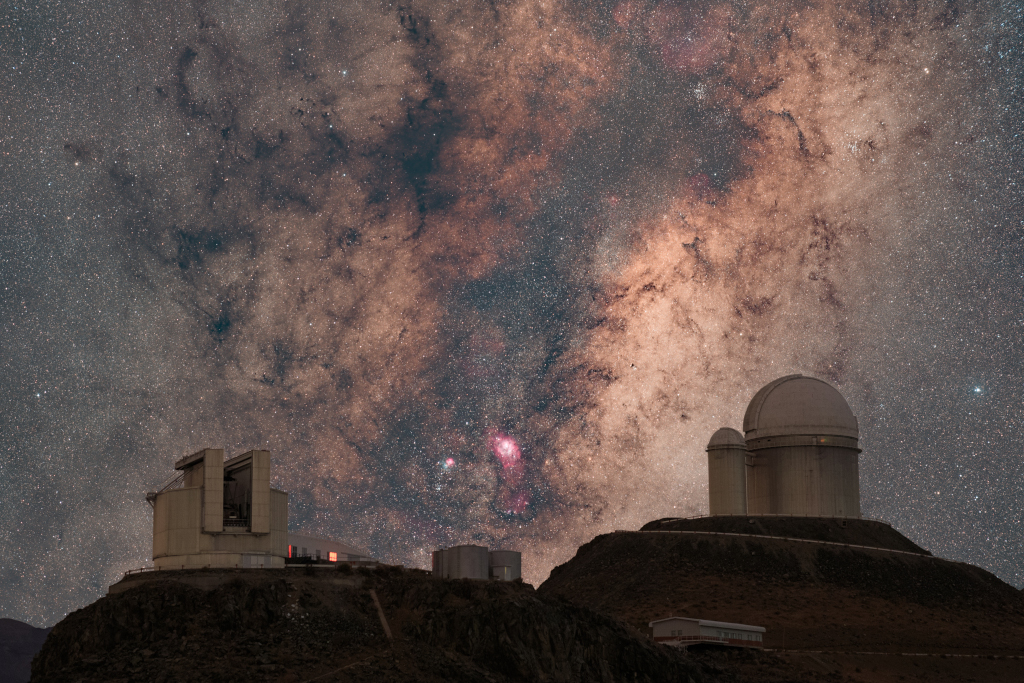 Hinter mehreren Teleskopkuppeln der Europäischen Südsternwarte ESO auf La Silla in Chile breitet sich die Milchstraße mit Dunkelwolken und leuchtenden Nebeln aus.